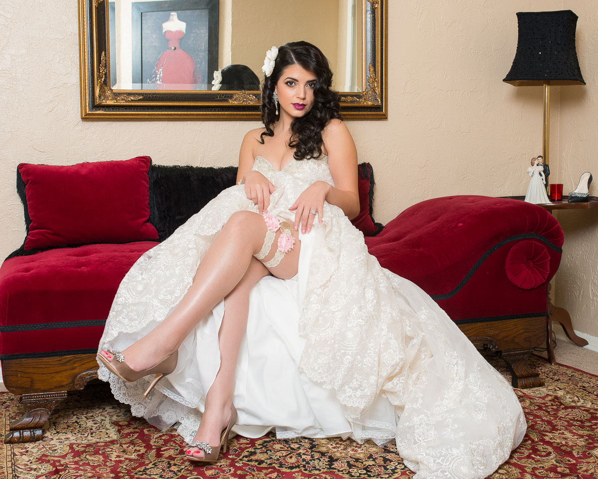 Bridal-1 A bride in a white gown sits elegantly on a red couch, legs crossed, exuding grace and anticipation.