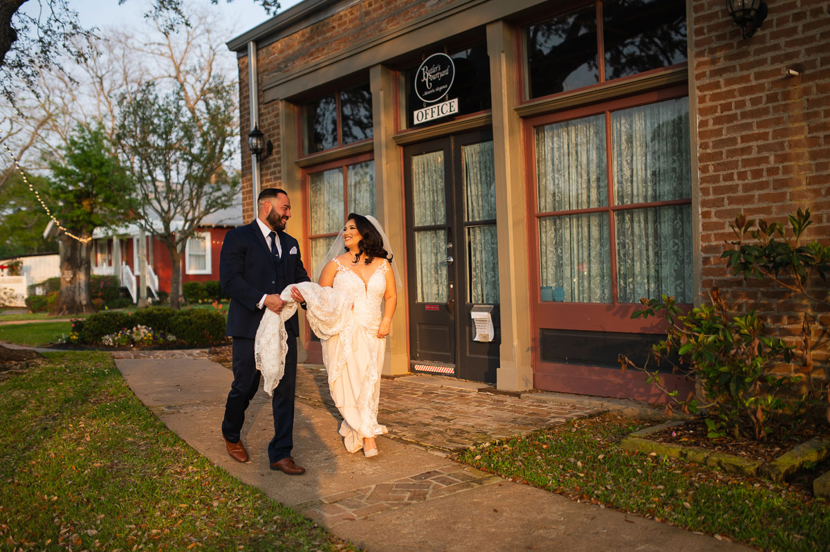 Destination-2-1 A bride and groom stroll hand in hand along a sidewalk beside a brick building, radiating joy on their wedding day.