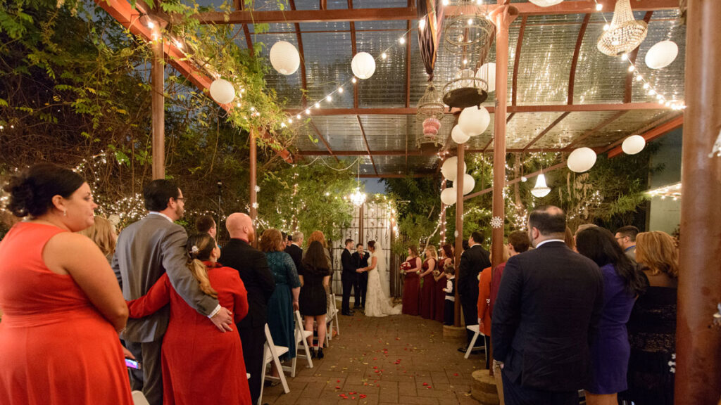 A wedding ceremony in a courtyard, featuring guests wearing elegant red dresses amidst floral decorations.