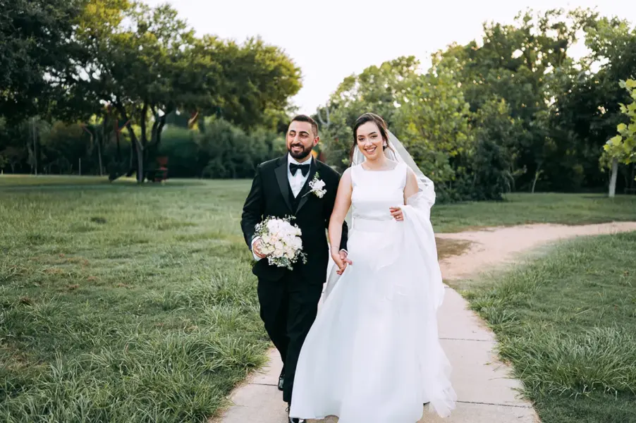 A bride and groom walk hand in hand along a garden path, with the groom carrying a bouquet and trees behind them.